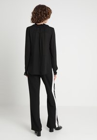 Musta pitkähihaine paita, jossa on frilla kauluksessa, yhdistettynä mustiin housuihin, joissa on valkoiset sivuraitat. Silkkinen kangas ja istuva malli.