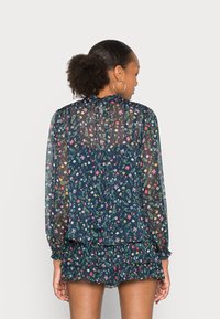 Blusa trasparente a stampa floreale in blu navy, con maniche lunghe, orlo arricciato e polsini elasticizzati. Il materiale sembra leggero e ha una finitura texture.