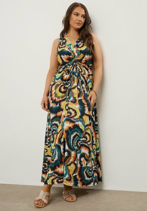 Kvinde iført en ærmeløs maxi-kjole med flerfarvet spiral mønster og en snoet frontdetalje samt sandaler, stående mod en ensfarvet væg.