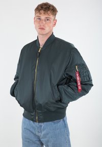 Tamnozeleni bomber jakna napravljena od glatkog najlona. Ima zlatni patentni zatvarač, rebraste rukave i donji deo, i crvenu dekorativnu oznaku na rukavu.
