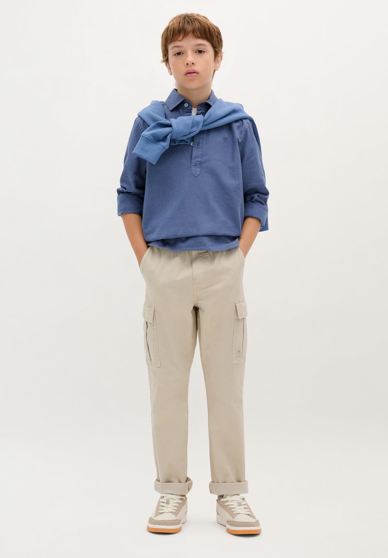 Blaues Langarmhemd mit Kragen, beige Cargo-Hosen und Sneakern. Pullover um den Hals gebunden. Taschen an den Hosen, lässiges Design.