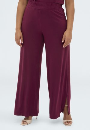Kobieta w bordowych, szerokich spodniach z elastycznym paskiem w talii i bocznymi rozcięciami, w połączeniu z beżowymi sandałami na obcasie.