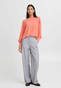Top leggero color corallo con maniche lunghe, abbinato a pantaloni grigi a gamba larga con un sottile motivo a righe e scarpe scure.