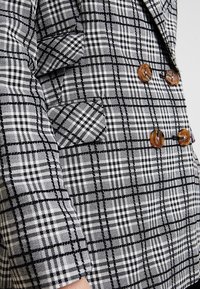 Kostkovaný blazer v černé a bílé barvě s hnědými knoflíky. Má klopový límec, dvouřadý design a detaily bočních kapes.
