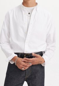 Osoba oblečená v bílé košili s knoflíky přes bílé tričko, tmavých džínách, černém opleteném pásku, stříbrných prstenech, náramku a dvou náhrdelnících s přívěsky.