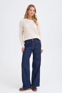 Pull en tricot beige à texture côtelée, associé à un jean large bleu foncé avec des poches avant et une fermeture à bouton. Chaussures marron.