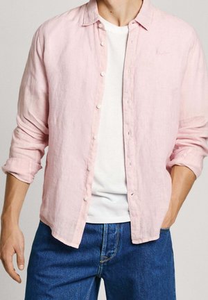 Lichtroze linnen shirt met een knoopsluiting aan de voorkant en een kraag, gelaagd over een wit T-shirt, gecombineerd met blauwe jeans.