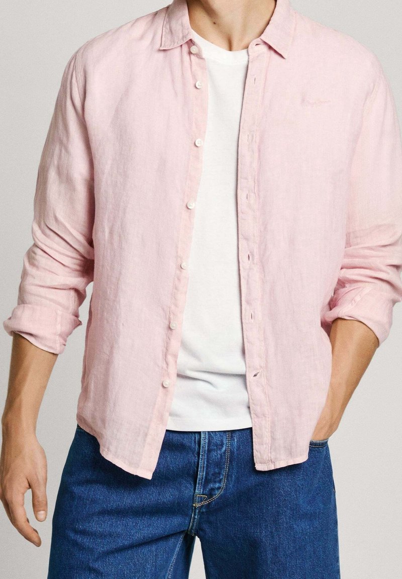 Lichtroze linnen shirt met een knoopsluiting aan de voorkant en een kraag, gelaagd over een wit T-shirt, gecombineerd met blauwe jeans.