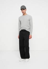 Άνδρας με γκρι μακρυμάνικο πουλόβερ, ταιριαστό γκρι καπέλο, μαύρο παντελόνι σε φαρδιά γραμμή και λευκά αθλητικά παπούτσια, στέκεται μπροστά σε λευκό εντελώς φόντο.