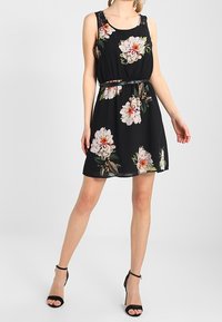 Schwarzes Blumenkleid mit ärmellosem Design; hat eine taillierte Taille und einen lockeren Rock. Auffällige weiße und rosa Blumen mit grünen Blättern.