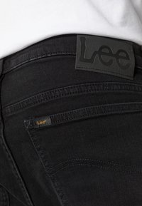 Svarta denimjeans har en strukturerad yta, med en stor svart läderlapp som visar "Lee" på midjebandet och en mindre etikett på fickan.