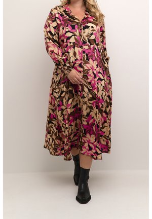 Kobieta ubrana w długą sukienkę na guziki z motywem kwiatowym w beżowe, różowe i brązowe liście, w czarnych botkach do kostki, stoi na tle jednolitej ściany.