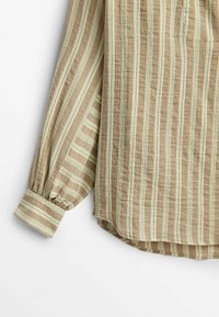 Chemise beige à manches longues avec rayures blanches verticales, poignets boutonnés et ourlet arrondi, tissu léger texturé.