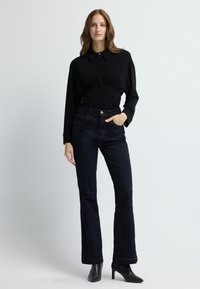 Musta pitkähihaine paita, jossa on istuva vyötärö ja kaulus, yhdistetty tummansinisiin leveälahkeisiin farkkuihin ja mustiin korkokenkiin.