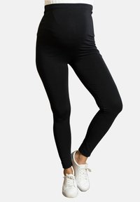 Leggings de maternidad negros con una cintura alta, textura suave y diseño ajustado, combinados con zapatos blancos sobre un fondo neutral.