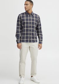 Cămașă cu mâneci lungi în carouri bleumarin și crem, cu închidere cu nasturi, purtată cu chinos bej deschis și adidași albi, având guler și manșete clasice.