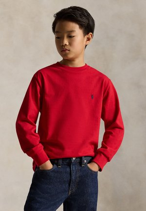 Garçon portant une chemise rouge à manches longues et un jean bleu, les mains dans les poches, regardant légèrement vers le bas et sur le côté.