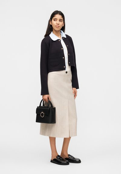 Γυναίκα που φοράει μαύρο cropped σακάκι, ανοιχτό γαλάζιο πουκάμισο με γιακά, μπεζ midi φούστα, μαύρα loafers και κρατάει μαύρη τσάντα σε λευκό φόντο.