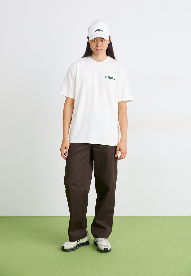 T-shirt bianco a maniche corte con testo verde, pantaloni marroni larghi e un cappellino bianco con testo nero. La calzatura presenta una suola testurizzata.