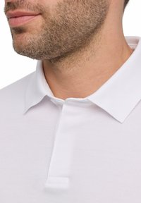 Witte polo met een zachte textuur, klassieke kraag en details van de voorste sluiting, gedragen op een deels zichtbare nek en kaaklijn.