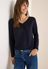 Marine blauwe langekam, met een V-hals, met geplooide details op de borst. Gecombineerd met licht beschadigde blauwe jeans.