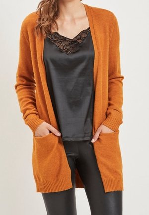 Femme portant un long cardigan orange brûlé avec des poches avant, par-dessus un haut en satin noir orné de dentelle, et un pantalon en cuir noir.