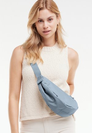 Giovane donna con capelli biondi che indossa un top senza maniche beige a maglia e una borsa a tracolla azzurro chiaro.