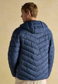 Marineblå pufferjakke med hætte. Har et chevron-quiltet design med vandrette linjer. Blød, glat tekstur. Holdbart materiale.