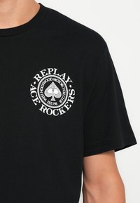 Camiseta de algodón negra con un gráfico circular blanco que destaca el texto "Replay" y "Ace Rockers", resaltando un diseño de trébol. Manga corta.