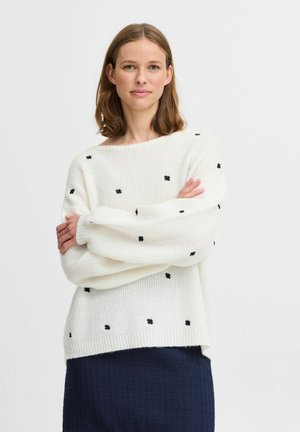 Maglione bianco lavorato a maglia con motivi floreali neri. Collo a barca ampio, maniche lunghe e texture a coste. Abbinato a una gonna texturizzata blu scuro.