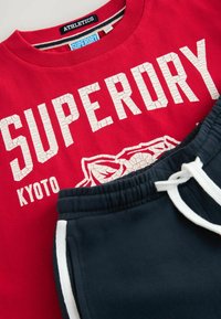 Červená mikina s bílým prasklým textem "SUPERDRY KYOTO" a obrysem grafiky. Námořnické tepláky s bílými postranními pruhy a šňůrkou na stažení.