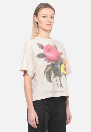 Donna bionda che indossa una maglietta beige oversize con una grande stampa floreale rosa e gialla, abbinata a pantaloni neri, in piedi davanti a uno sfondo bianco.