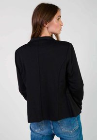 Svart blazer med en strukturerad design, slagkrage, slät yta och skräddarsydd passform, sett från baksidan.