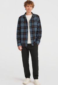 Camisa de franela a cuadros negro y azul con mangas largas, cierre de botones en la parte delantera y dos bolsillos en el pecho, combinada con pantalones negros y zapatillas blancas.