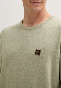 Jasnozielony sweter z okrągłym dekoltem, wykonany z fakturowanej tkaniny, z małą kwadratową łatą z logo w czerni i pomarańczu na klatce piersiowej.