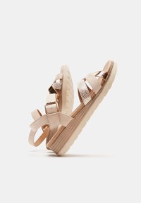 Sandales rose pâle avec des brides à la cheville, un accent doré texturé et une semelle robuste à motifs. Elles sont dotées de brides croisées et de boucles réglables.