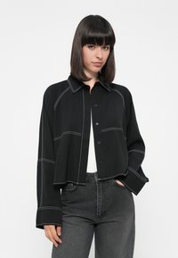Black cropped shirt with contrasting white stitching, button-front closure, and a collar. Paired with high-waisted gray jeans.