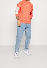 Korallfärgad sweatshirt med logotyp, texturerade ärmar i mjuk mintgrön färg, kombinerad med ljusblå jeans med raka ben och vita sneakers.