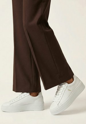 Witte lage sneakers gedragen met wijde bruine broekspijpen op een lichte achtergrond, waarbij één voet iets is opgetild.