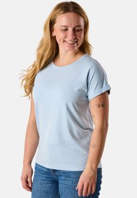 T-shirt di cotone azzurro chiaro con maniche arrotolate, scollatura rotonda e una texture liscia. Indossato con jeans blu, caratterizzati da dettagli di tatuaggi sottili.