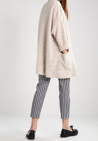 Beige übergroßer Pullover mit umgeschlagenen Ärmeln und seitlichen Taschen, kombiniert mit schwarz-weißen Karo Slim-Fit-Hosen und schwarzen Slippern.