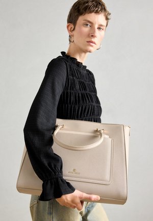 Borsa a tracolla beige con manico superiore, finitura testurizzata, tasca frontale e accenti dorati. Indossata da un modello con una blusa nera con volant.