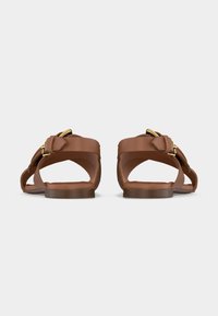 Καφέ δερμάτινα σανδάλια, επίπεδου τύπου, με λουράκια και αγκράφες σε χρυσό τόνο, σε απόψη από πίσω σε απλό λευκό φόντο.