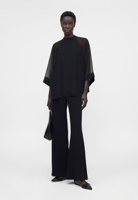 Femme portant un top noir à manches transparentes, pantalon large, chaussures pointues, tenant un sac à main noir, debout sur fond blanc.