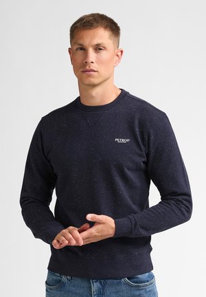 Mann trägt marineblauen gesprenkelten Pullover mit "Petrol Industries" Logo, steht mit verschränkten Händen, neutralem Gesichtsausdruck, einfarbigem hellem Hintergrund.