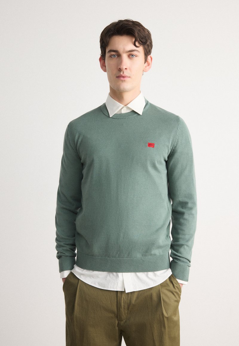 Pull en maille vert avec col rond, poignets côtelés et ourlet côtelé. Présente une petite patch logo rouge sur la poitrine. Porté par-dessus une chemise à col.