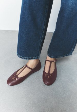Pieds portant des ballerines rouges foncées à bride en T avec découpes décoratives, associées à un jean bleu à jambes larges, debout sur un sol gris clair.