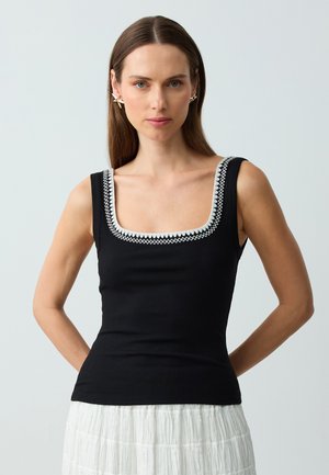 Schwarzes geripptes Tanktop mit quadratischem Ausschnitt und weißen gestickten Details am Rand, getragen mit einem weißen strukturierten Rock.