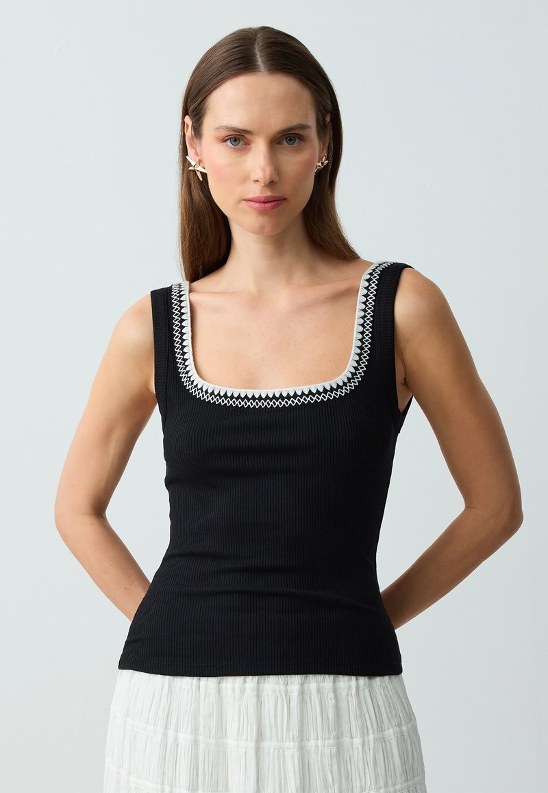 Top in cotone nero a costine con scollatura quadrata e dettagli ricamati bianchi lungo il bordo, indossato con una gonna bianca a trama.