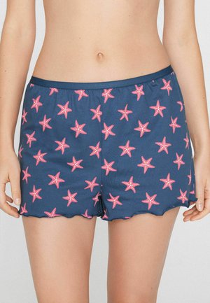 Marineblaue Shorts mit pinkem Seesternmuster, getragen von einer stehenden Person, die den Bauch zeigt und die Hände entspannt an den Seiten hält.